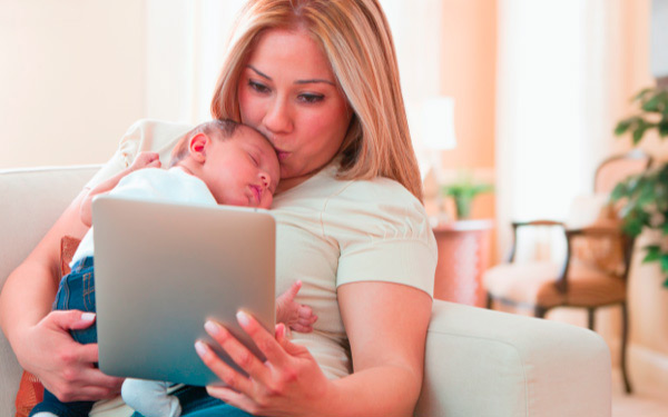 Woman cuddling and kissing a baby while looking at a tablet