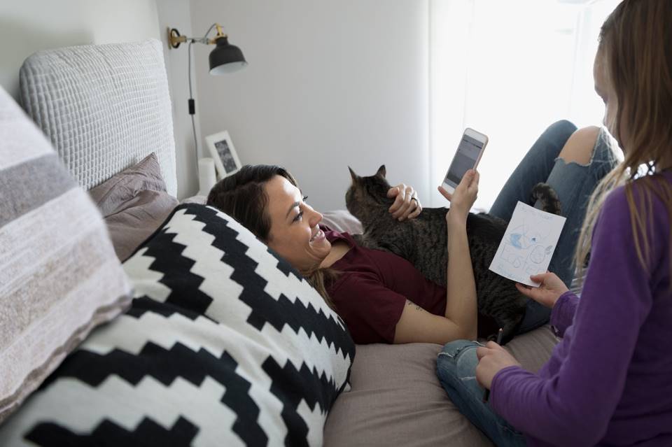 A woman laying on the bed, holding her phone and cuddling her cat. A child sits close by showing a drawing they have created.