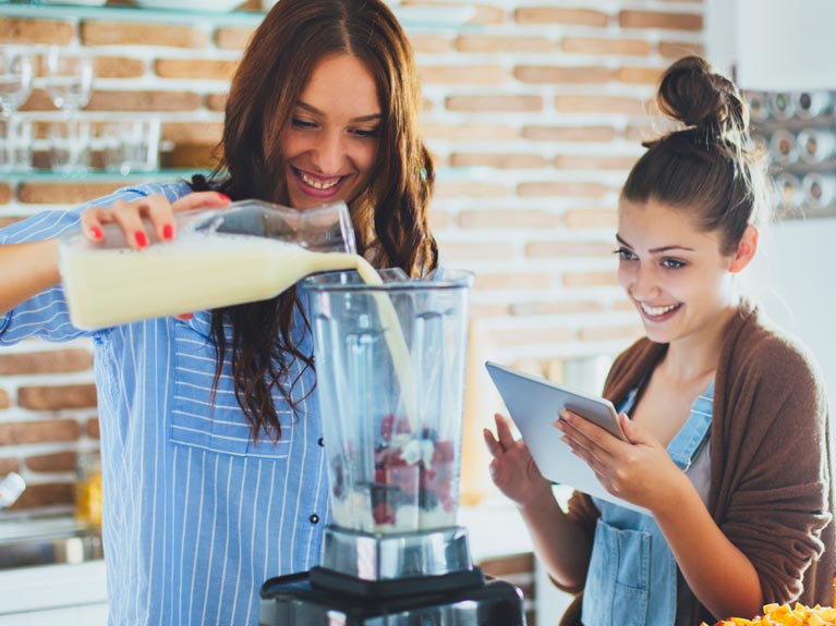 Two womwn. One pouring milk into a blender.