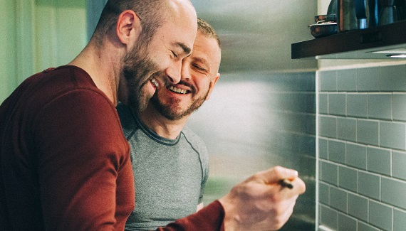 Two men in a kitchen smiling and about to taste something.