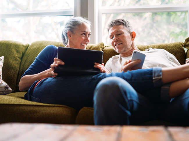 Father on a laptop device and son on a mobile device, in discussion on a sofa.