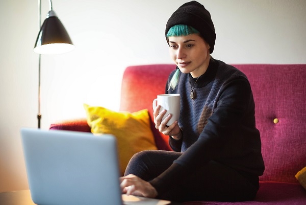 Image of a lady on her couch, holding a cup and looking at her laptop.